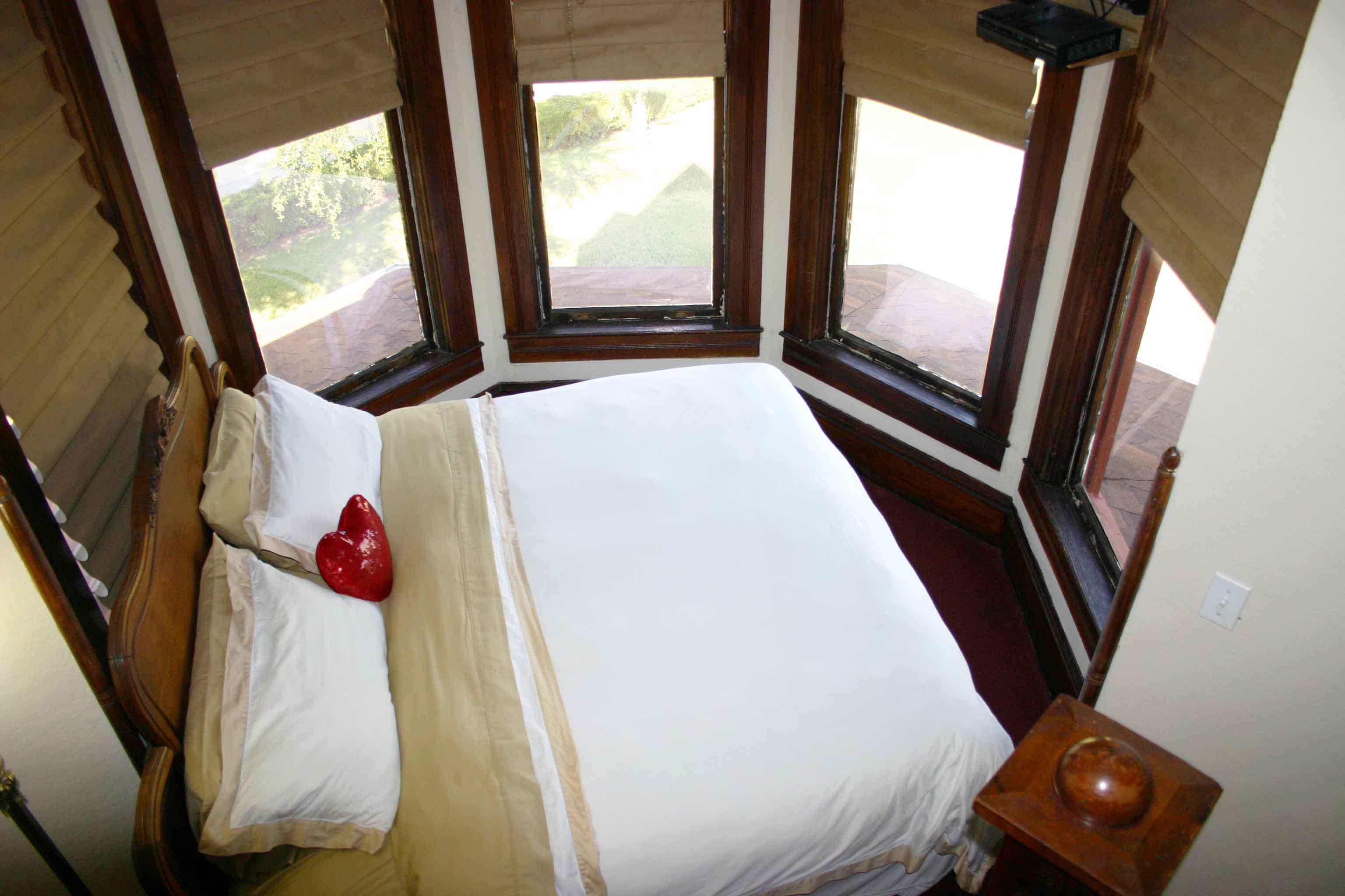 A bed with white sheets and pillows in front of two windows.